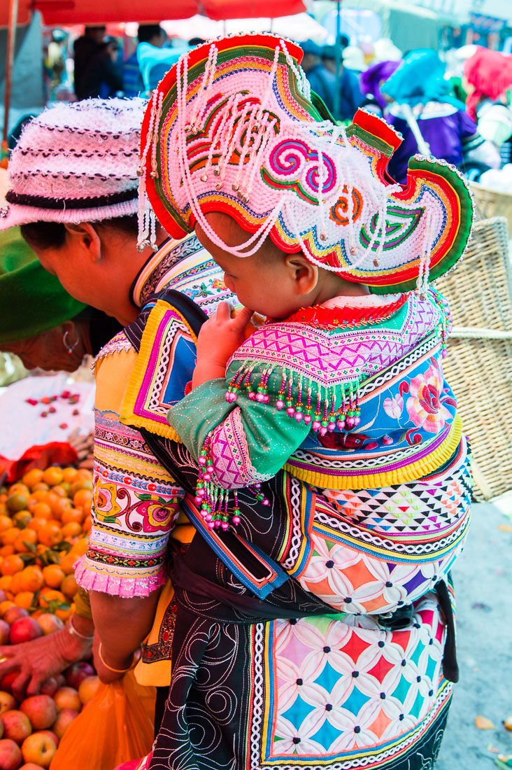 A mother from the Yi ethnic minority people carrying her baby, wearing traditional costume at the Sha La Tuo market, Yang Yang county, Yunnan Procince, China, Asia. Nikon D4, 24-120mm, f/ 4.0, VR.