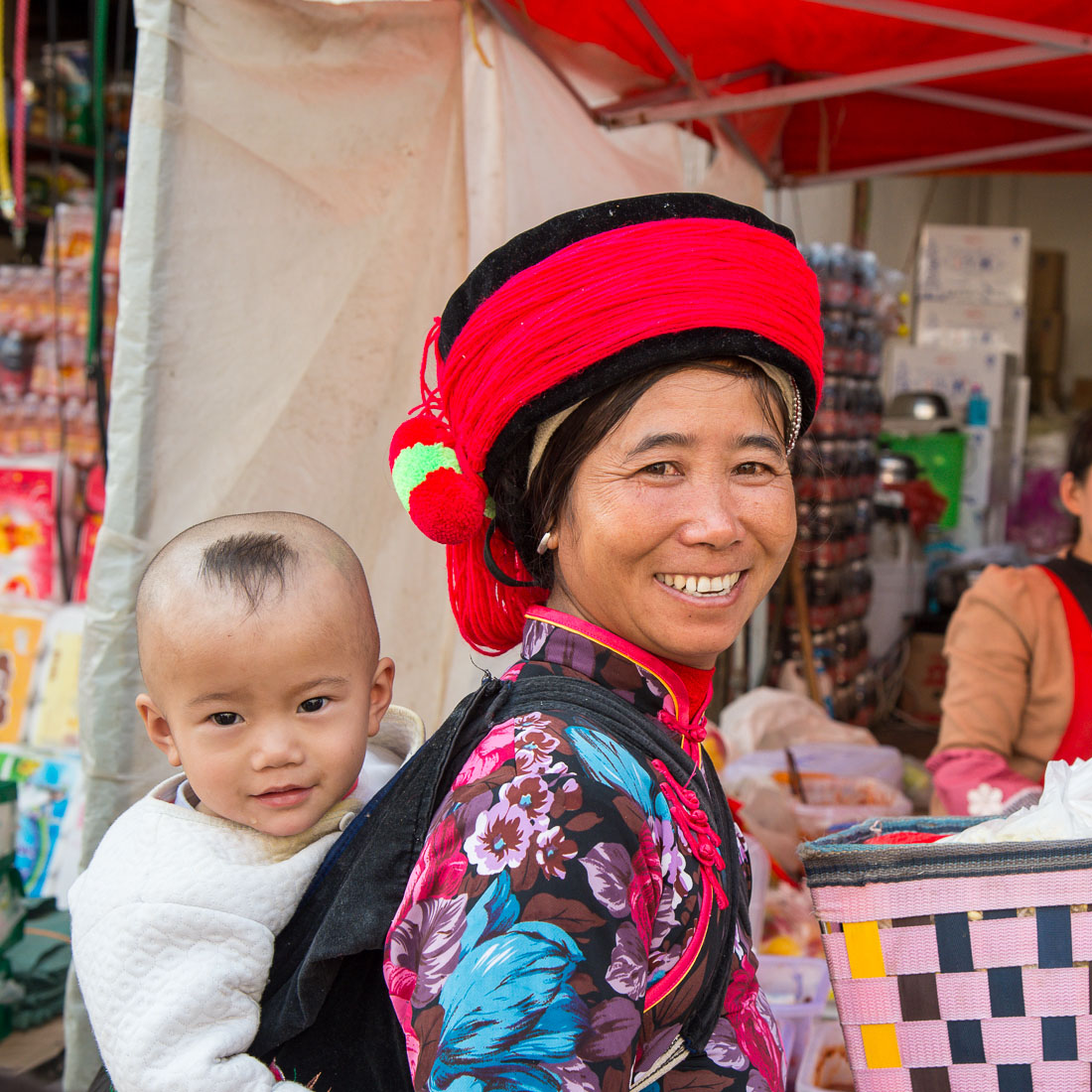 A joyful mother from the Ha Ni ethnic minority people carrying her baby, wearing traditional costume at the Ping Hi town market, Lv Chun county, Yunnan Procince, China, Asia. Nikon D4, 24-120mm, f/ 4.0, VR.
