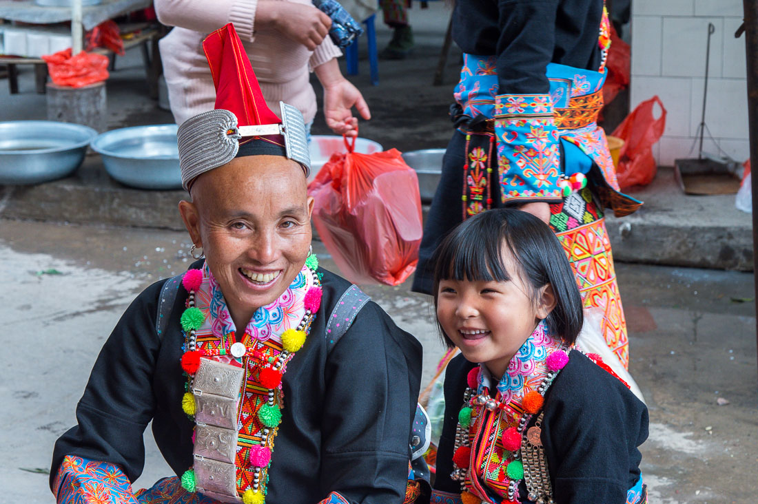 A young grandma with her granddaughter, both showing a large happy smile, wearing the traditional costumes of the Yao ethnic minority people at the Jin Ping market, Yunnan Province, China, Asia. Nikon D4, 24-120mm, f/4.0, VR