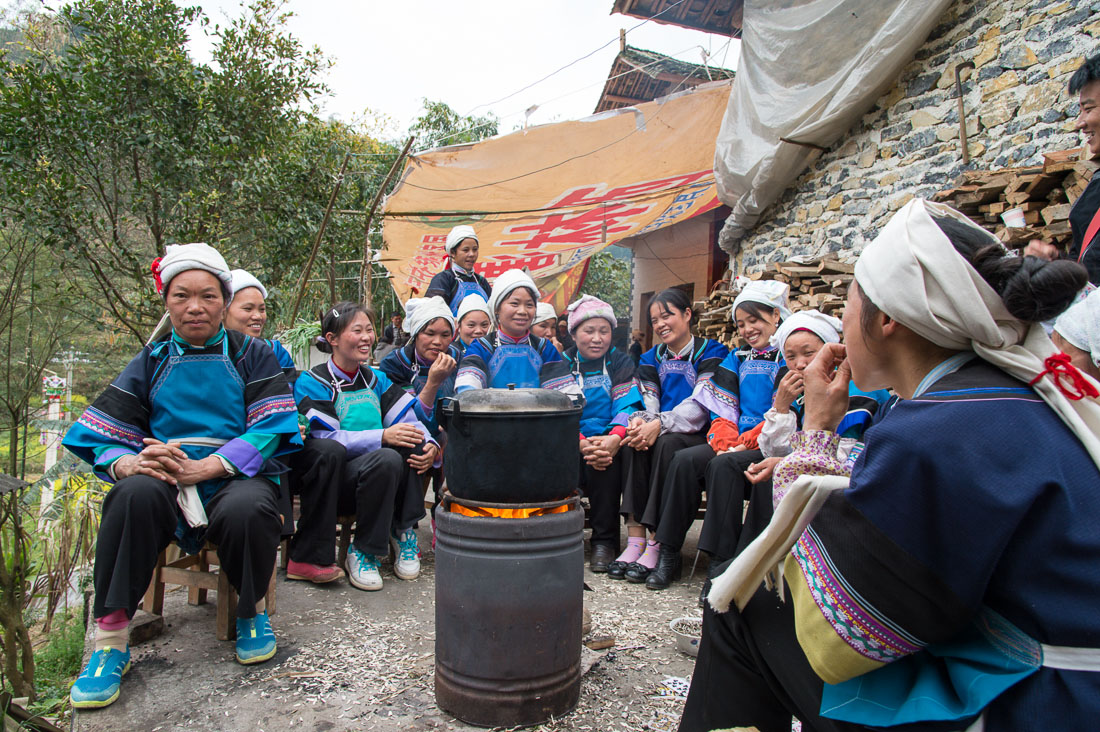 Women from the Bu Yi ethnic minority people, in traditional costume, attending a funeral, gathering next to a cooking fire, eating sunflower seeds. La Zhe village, Luo Ping County, Yunnan Province, China, Asia. Nikon D4, 24-120mm, f/4.0, VR