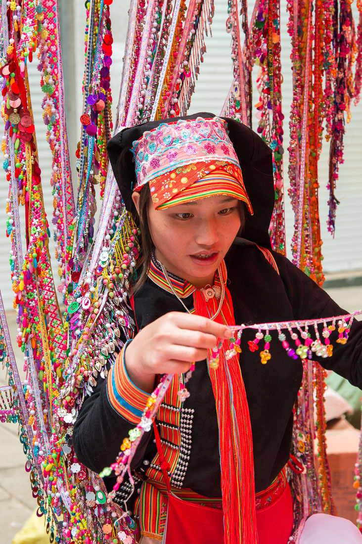 Young woman from the Yao ethnic minority people wearing the traditional costume at Jin Shui He market buying colorful ornaments. Jin Ping County, Yunnan Province, China, Asia. Nikon D4, 70-200mm, f/2.8, VR II