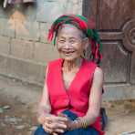 A very happy and smiling old woman from the Khamu people ethnic minority wearing her traditional costume, sitting in front oh her house; visible red stains on her teeth and the mouth area due to the chewing of paan (areca nuts and betel leaves). Lao PDR, Laos, Indochina, South East Asia