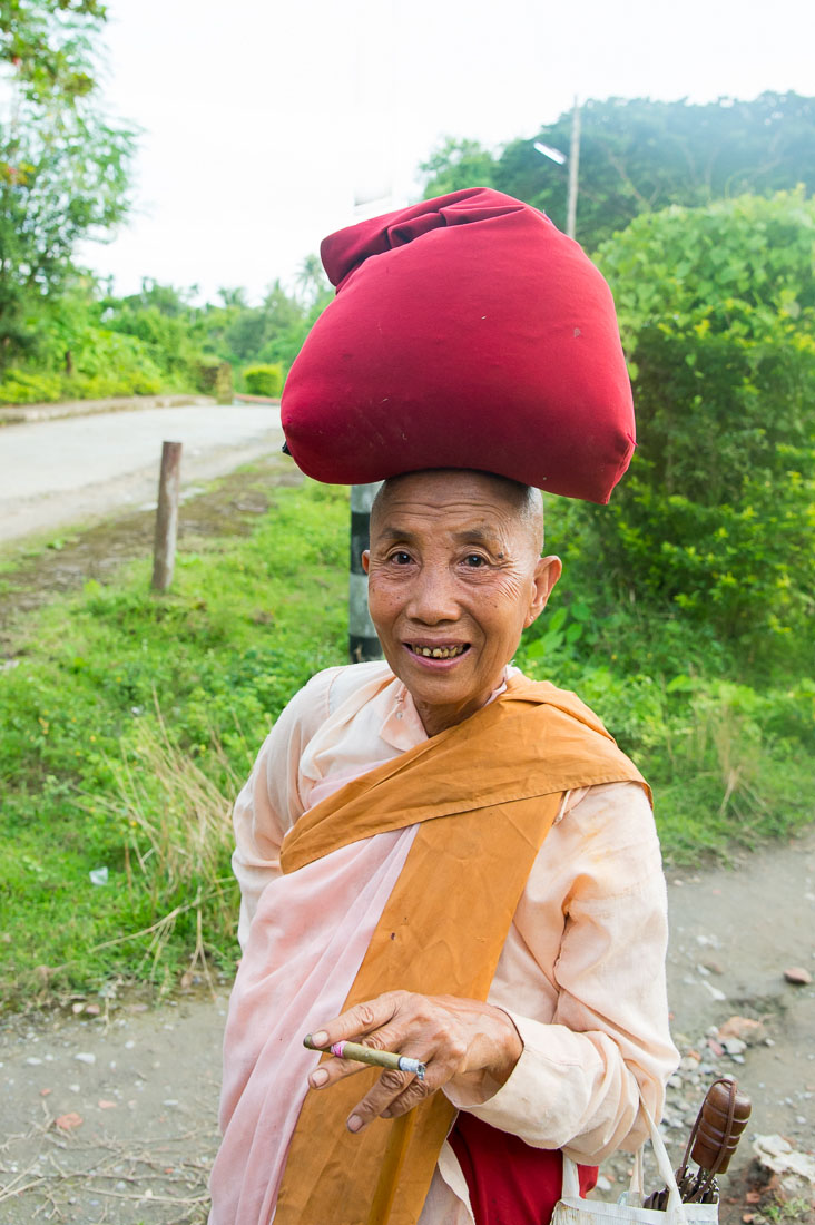 A happy old Buddhist nun smoking a cigar, walking back to the convent, carrying on her head alms of rice. Mrauk U Village, Rakhine State, Myanmar, Indochina, South East Asia.