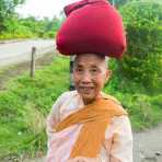 A happy old Buddhist nun smoking a cigar, walking back to the convent, carrying on her head alms of rice. Mrauk U Village, Rakhine State, Myanmar, Indochina, South East Asia.