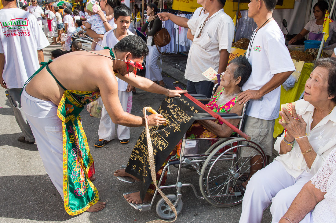 Psychic medium follower of the Bang New Shrine, with long needles pierced through his ears, offering spiritual blessing to a sick woman in a wheel chair, during a street procession in the annual Chinese vegetarian festival. Phuket, Kingdom of Thailand, Indochina, South East Asia